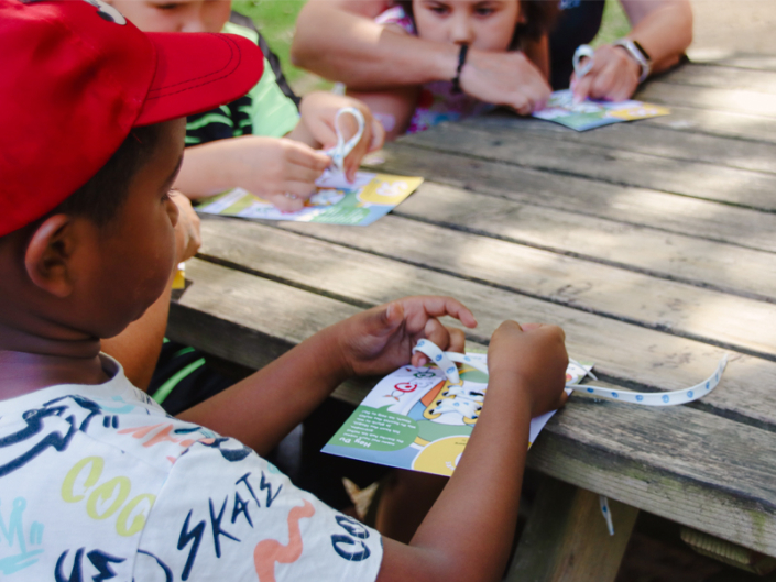 Kinder basteln an einem Holztisch im Freien, während sie mit bunten Papierkarten und einem Maßband arbeiten. Ein Junge in einem roten Hut konzentriert sich auf seine Aufgabe, während andere Kinder ebenfalls aktiv teilnehmen.