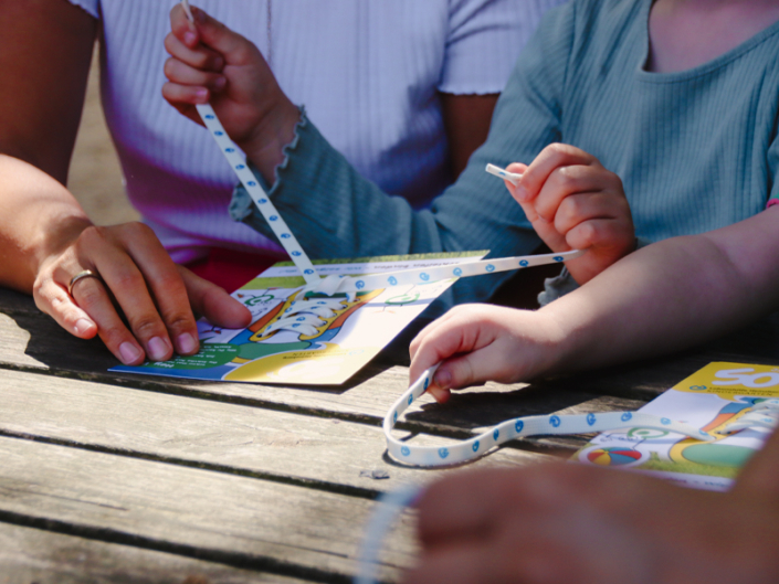 Hände von Erwachsenen und Kindern, die an einem Tisch sitzen und an Bastelmaterialien arbeiten. Auf dem Tisch liegen bunte Bastelblätter und Schnüre, die für kreative Aktivitäten verwendet werden.