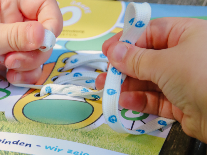 Hände binden weiße Schnürsenkel mit blauen Mustern auf einem bunten Spieluntergrund, der Schuhe darstellt.