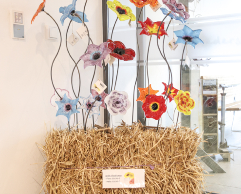 Bunte Blumen stehen in einem Heuballen. Hinter den Blumen ist ein Zaun und ein kleines Häuschen zu sehen.