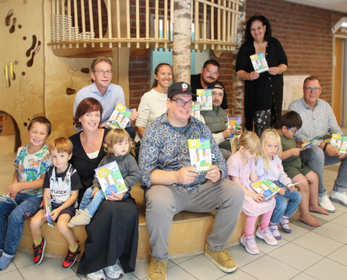 Viele Kinder und Erwachsene sitzen zusammen. Sie halten Bücher in der Hand.
