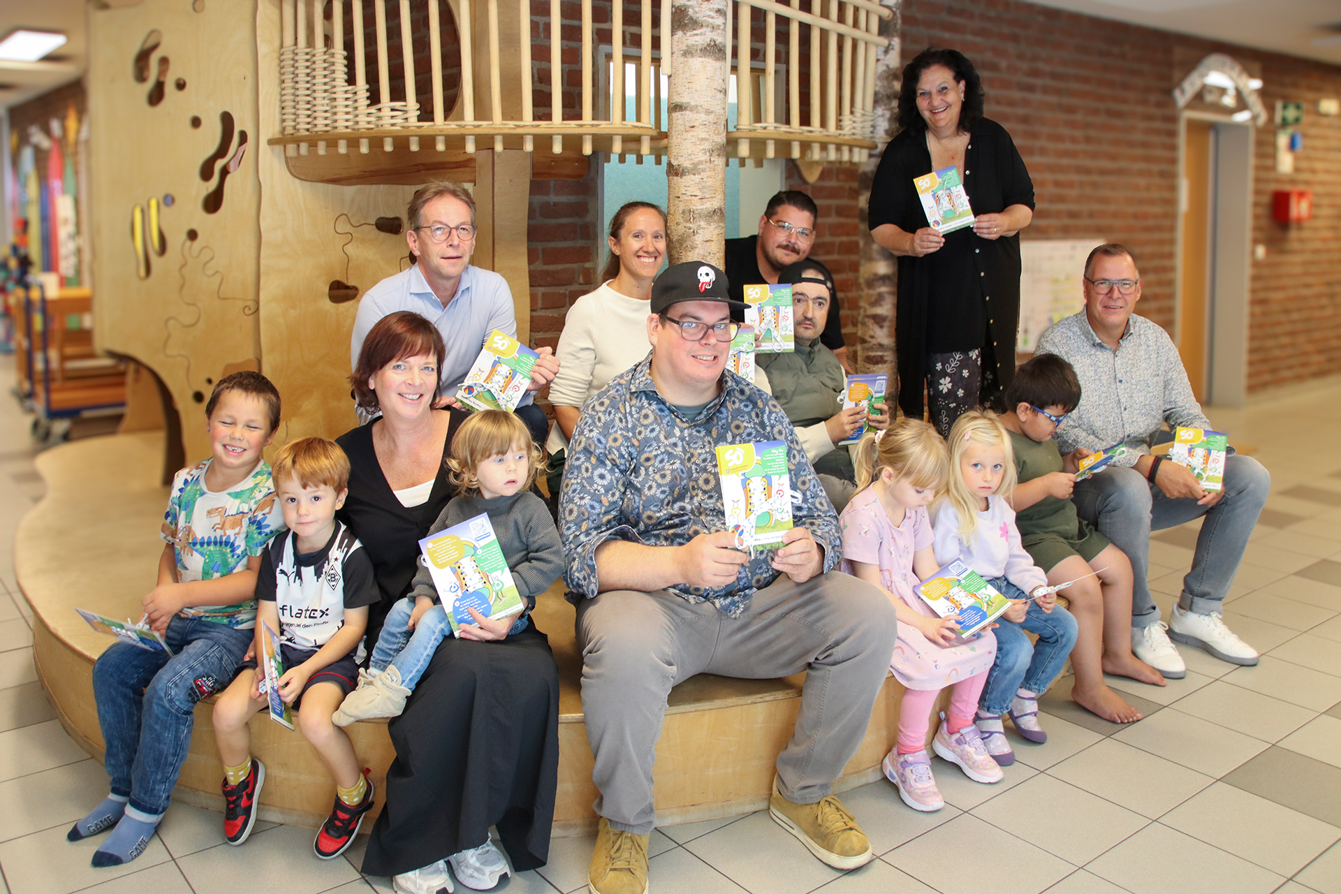 Viele Kinder und Erwachsene sitzen zusammen. Sie halten Bücher in der Hand.