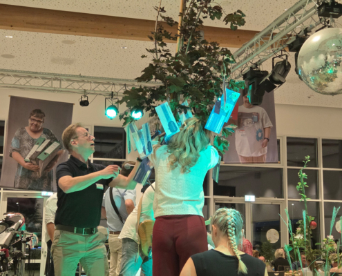 Eine Frau steht auf einer Bühne. Sie hat lange Haare. Ein Mann hilft ihr. Es gibt viel Technik im Hintergrund.