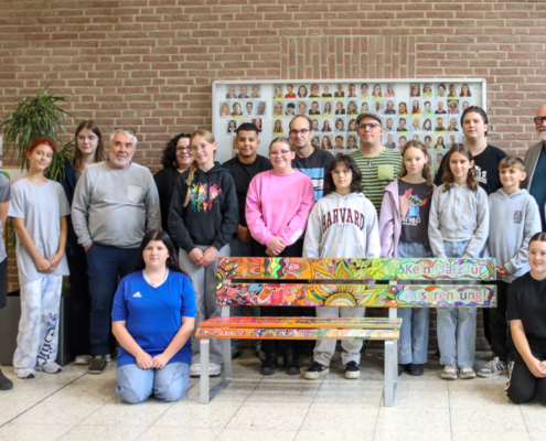 Eine Gruppe Menschen steht vor einem Gebäude. Manche sitzen auf einer bunten Bank.