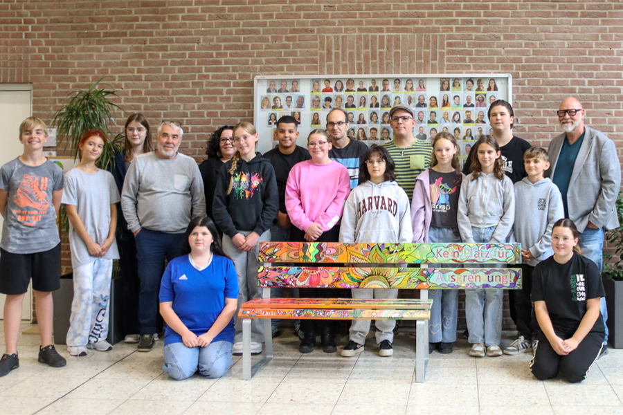 Eine Gruppe Menschen steht vor einem Gebäude. Manche sitzen auf einer bunten Bank.