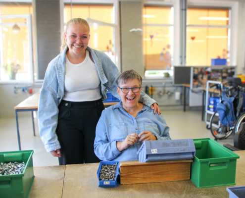 Zwei Frauen lächeln in einer Werkstattumgebung, während sie mit Schrauben und Werkzeugen arbeiten. Die jüngere Frau steht hinter der älteren, die sitzt und einen Kasten mit Schrauben hält. Im Hintergrund sind Tische und Arbeitsmaterialien sichtbar, was auf eine produktive Atmosphäre hinweist.