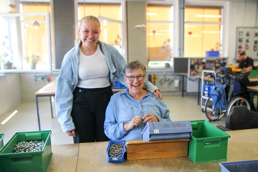 Zwei Frauen lächeln in einer Werkstattumgebung, während sie mit Schrauben und Werkzeugen arbeiten. Die jüngere Frau steht hinter der älteren, die sitzt und einen Kasten mit Schrauben hält. Im Hintergrund sind Tische und Arbeitsmaterialien sichtbar, was auf eine produktive Atmosphäre hinweist.