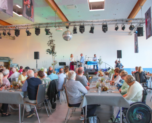 Viele Menschen sitzen an Tischen. Eine Frau spricht auf einer Bühne. Es gibt eine Barrierefreie Stellfläche für eine Rollstuhlfahrer Person.