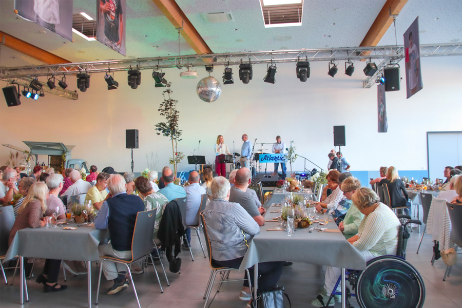 Viele Menschen sitzen an Tischen. Eine Frau spricht auf einer Bühne. Es gibt eine Barrierefreie Stellfläche für eine Rollstuhlfahrer Person.
