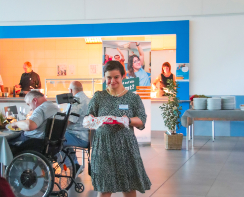 Eine Frau steht in einem hellen Raum. Sie trägt ein Kleid und Sandalen. Im Hintergrund sitzen Menschen und essen.