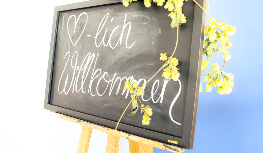 Schwarzes Schild mit der handgeschriebenen Begrüßung "Herzlich Willkommen", umgeben von Hopfenblüten, auf einem Holzständer platziert.