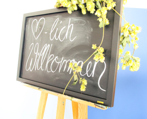 Schwarzes Schild mit der handgeschriebenen Begrüßung "Herzlich Willkommen", umgeben von Hopfenblüten, auf einem Holzständer platziert.