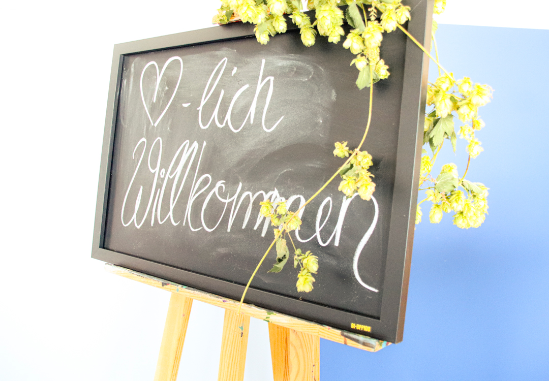 Schwarzes Schild mit der handgeschriebenen Begrüßung "Herzlich Willkommen", umgeben von Hopfenblüten, auf einem Holzständer platziert.