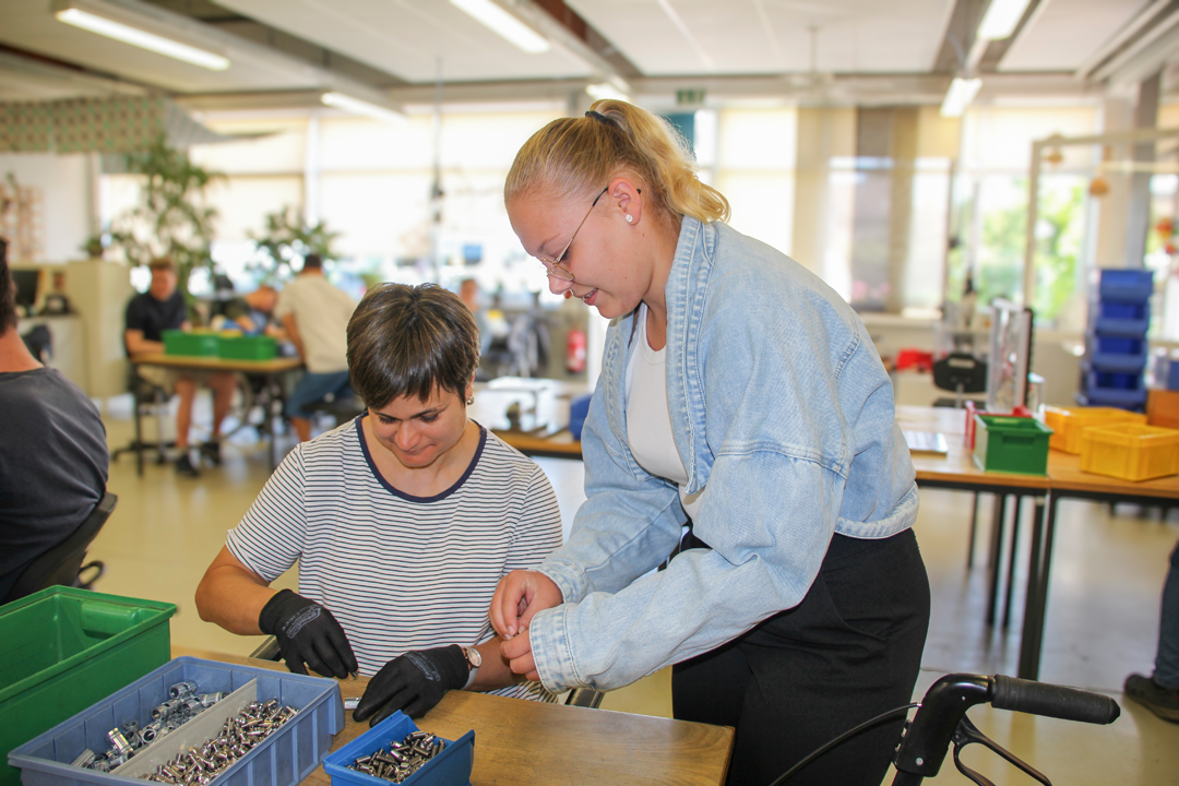 Zwei Frauen arbeiten in einer Werkstatt, während sie gemeinsam an einem Projekt mit Schrauben und Werkzeugen arbeiten. Im Hintergrund sind weitere Personen und Arbeitsplätze zu sehen, die eine produktive Umgebung schaffen.
