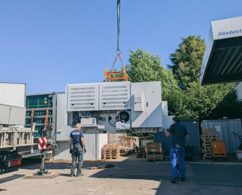 Ein Kran hebt eine große Kiste. Zwei Männer stehen daneben. Es ist ein Lager oder eine Fabrik.