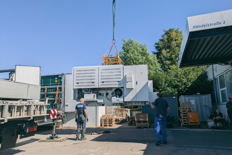 Ein Kran hebt eine große Kiste. Zwei Männer stehen daneben. Es ist ein Lager oder eine Fabrik.