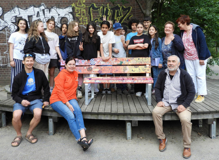 Menschen sitzen auf einer Bank im Park. Es sind verschiedene Personen, darunter Kinder und Erwachsene.