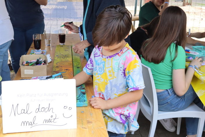 Kinder sitzen an einem Tisch und malen mit Buntstiften. Es gibt Papier und Kisten mit Dingen zum Basteln.