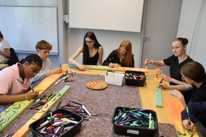 Kinder sitzen an einem Tisch und malen mit Buntstiften auf Holzbretter.