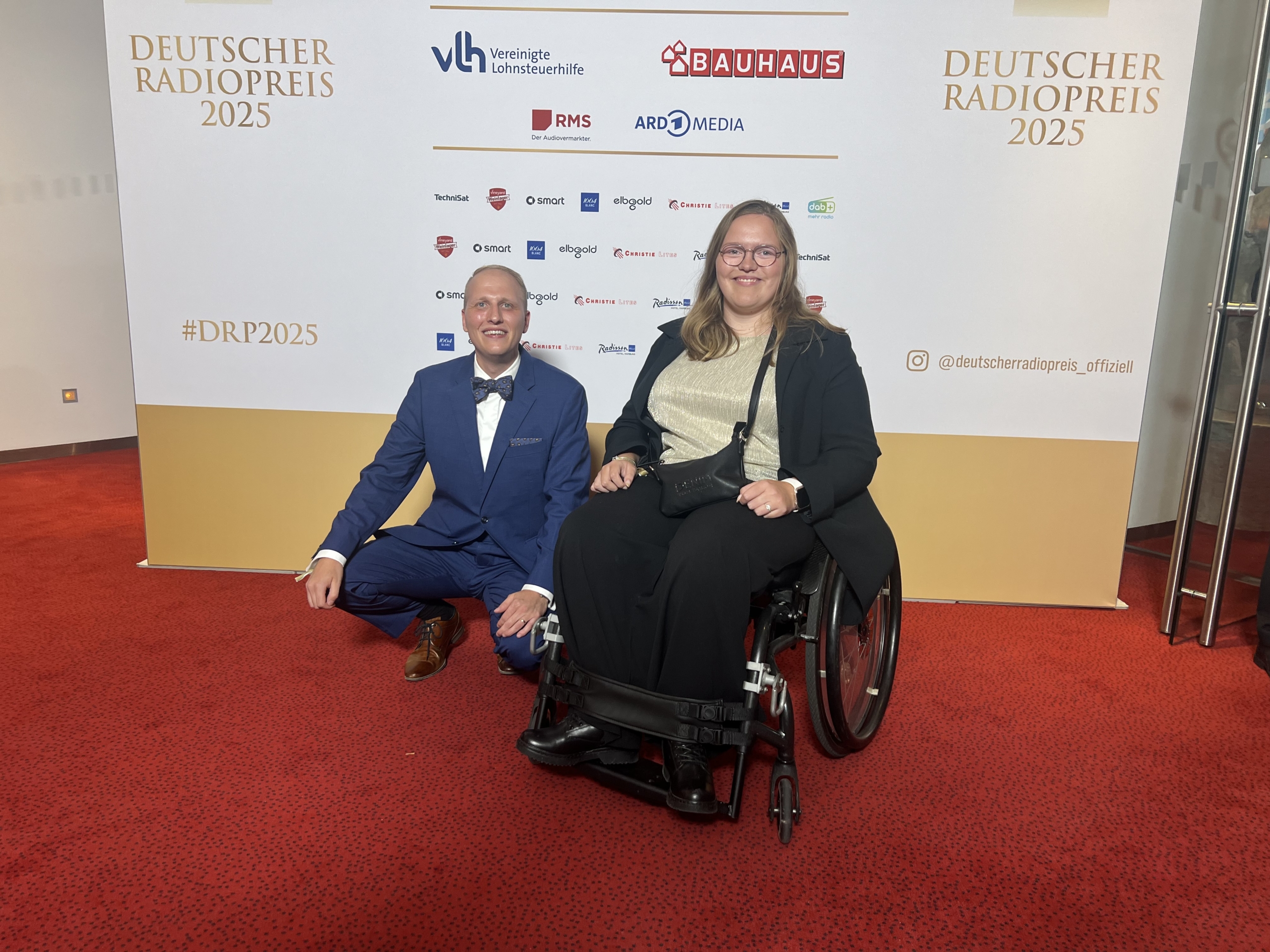Zwei Personen sitzen auf einem roten Teppich. Ein Mann trägt einen Anzug, eine Frau sitzt im Rollstuhl.