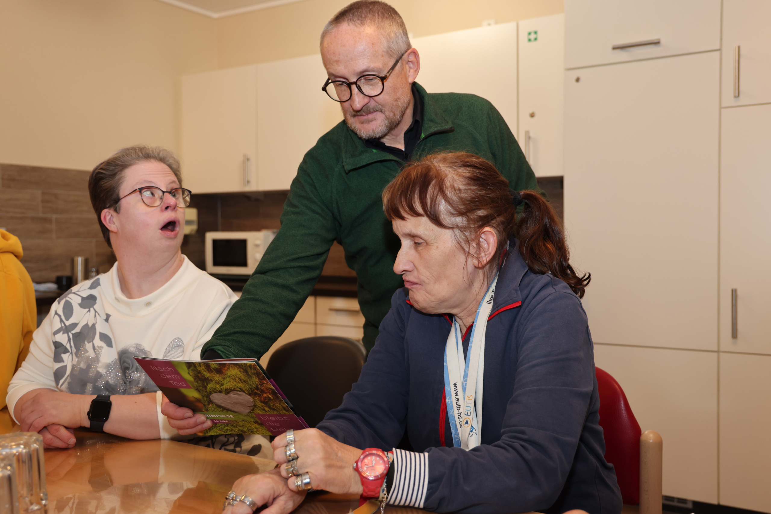Zwei Personen schauen ein Buch an. Der Mann hat eine grüne Jacke an. Die Frau trägt ein blaues Shirt.