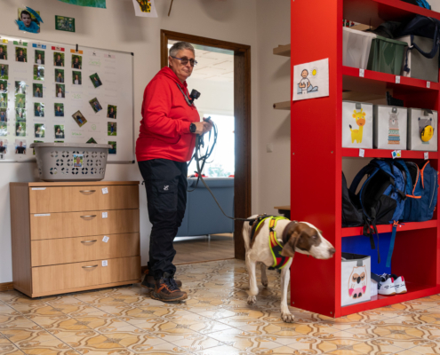 Ein Mann steht mit seinem Hund in einem Zimmer. Der Mann trägt eine rote Jacke. Der Hund hat einen roten Halsband.