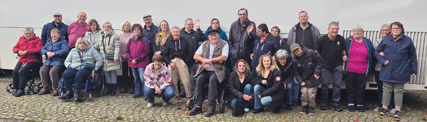 Eine Gruppe älterer Menschen steht vor einer Mauer. Manche stehen, manche sitzen auf einer Bank.