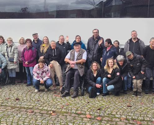Eine Gruppe älterer Menschen steht vor einer Mauer. Manche stehen, manche sitzen auf einer Bank.