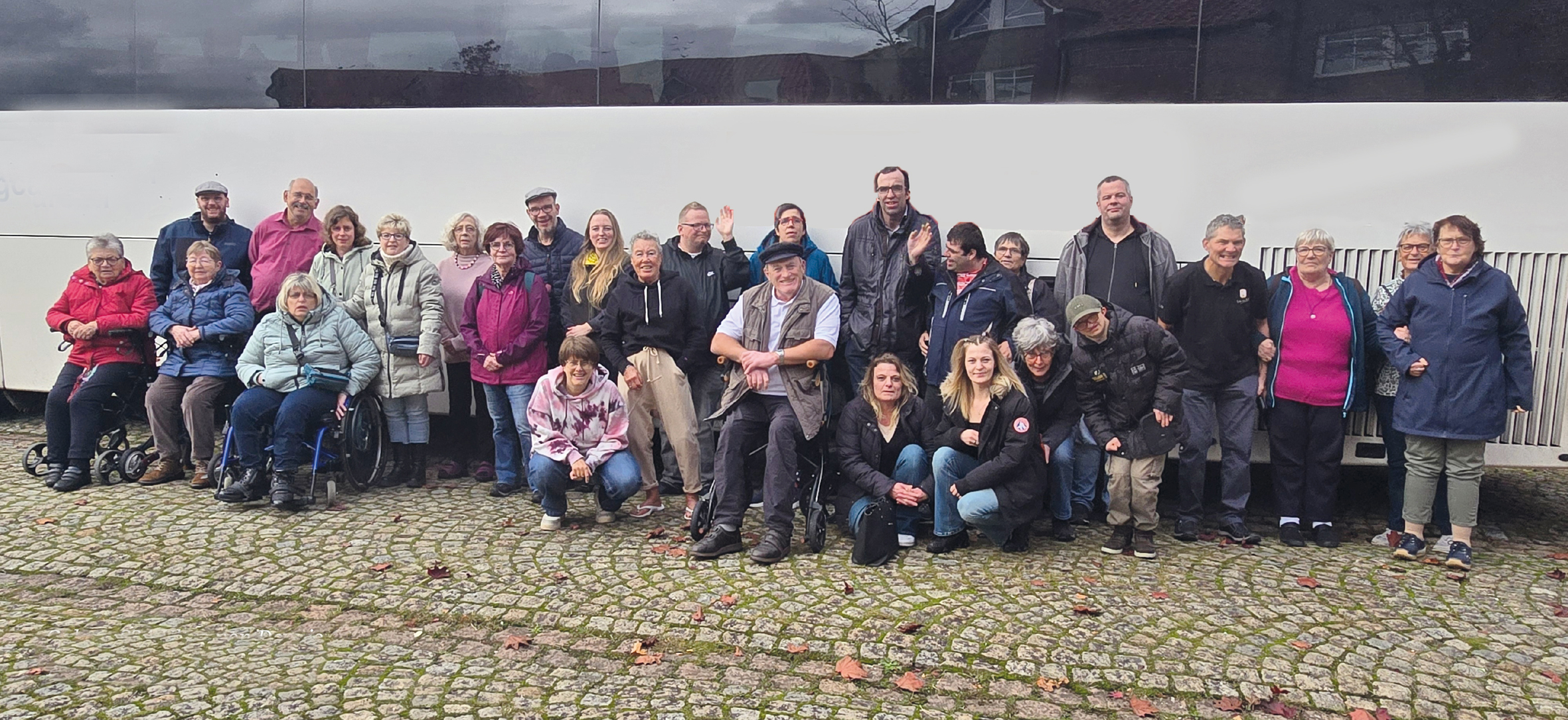 Eine Gruppe älterer Menschen steht vor einer Mauer. Manche stehen, manche sitzen auf einer Bank.