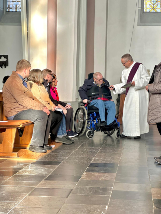 Menschen sitzen in einer Kirche. Ein Mann im weißen Gewand spricht. Ein Mann sitzt im Rollstuhl.