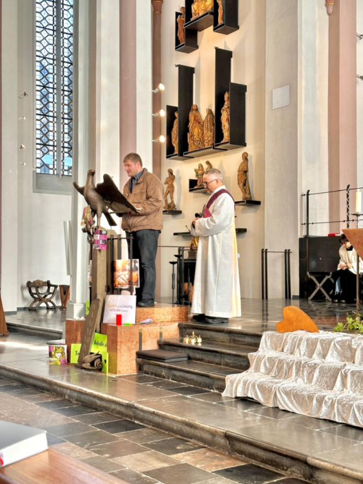Ein Mann in weißer Kleidung steht vor einem Altar. Er hält ein Buch in der Hand. Es stehen Figuren und Pflanzen davor.