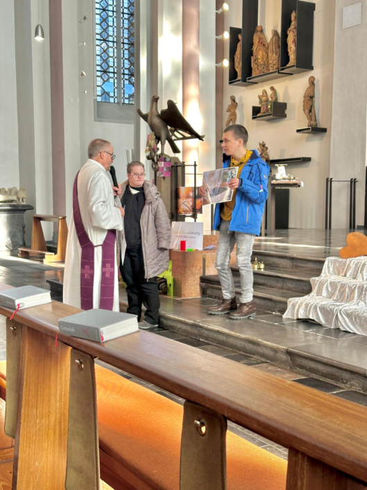 Drei Männer stehen in einer Kirche. Einer hat einen schwarzen Umhang an. Sie schauen auf ein Papier.