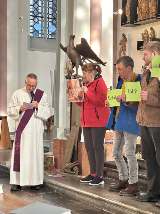 Ein Mann in weißer Kleidung hält ein Buch. Er steht in einem Raum mit Bildern und Figuren. Es sind noch andere Menschen im Raum.