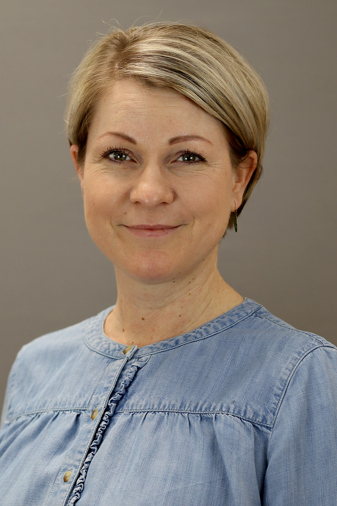 Eine Frau mit kurzen Haaren und Brille lächelt in die Kamera. Sie trägt ein blaues Hemd.