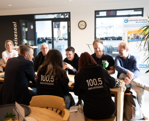 Eine Gruppe von Menschen sitzt an einem Tisch. Sie unterhalten sich und trinken Kaffee. Es sieht nach einem gemütlichen Treffen aus.