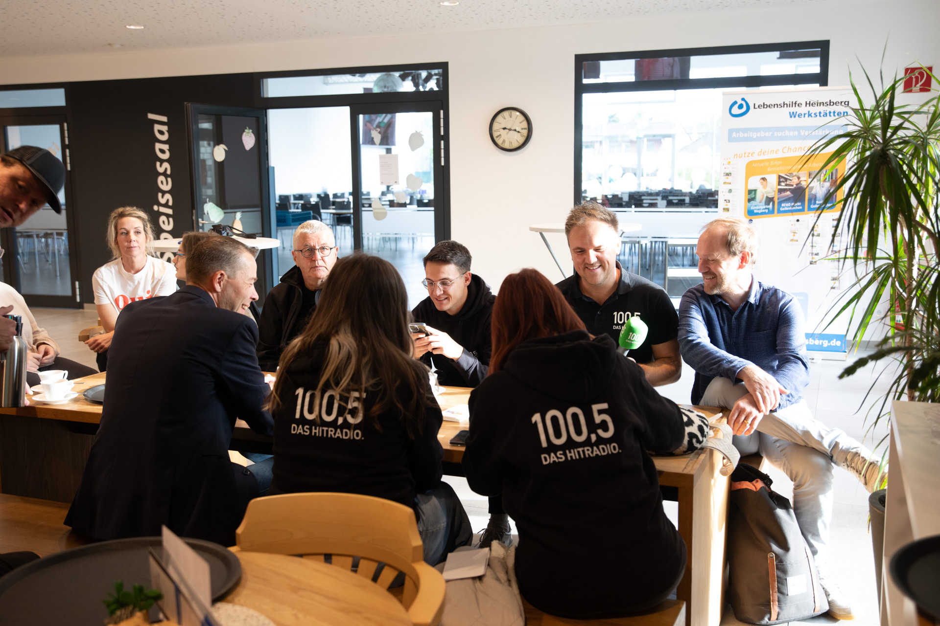 Eine Gruppe von Menschen sitzt an einem Tisch. Sie unterhalten sich und trinken Kaffee. Es sieht nach einem gemütlichen Treffen aus.
