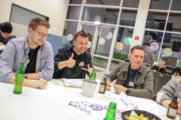 Drei Männer sitzen an einem Tisch. Sie trinken Bier und lächeln. Auf dem Tisch sind Gläser und Essen.