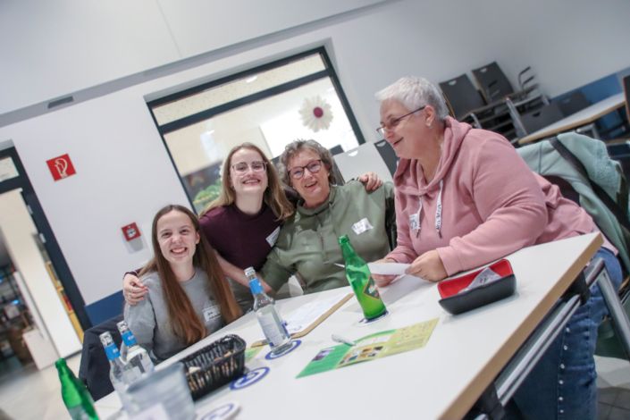 Zwei Frauen sitzen an einem Tisch. Sie schauen sich etwas an und lächeln. Auf dem Tisch liegen Papiere und Getränke.