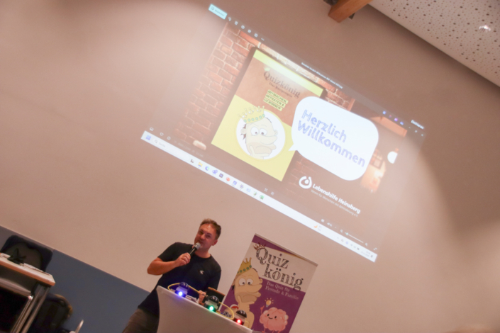 Ein Mann steht vor einer Leinwand und spricht. Hinter ihm sind viele kleine Lichter.