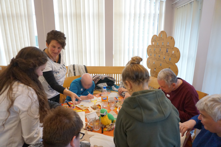 Menschen sitzen um einen Tisch. Sie basteln und lachen. Es gibt Papier, Farbe und andere Sachen zum Basteln.