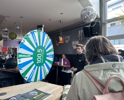 Zwei Personen sitzen in einem Café. Ein Mann schaut nach draußen. Es gibt Tische und Stühle.