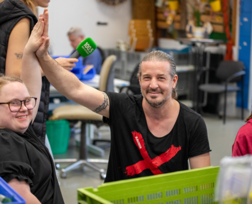 Ein Mann lacht und winkt. Er hat kurze, dunkle Haare und trägt ein schwarzes Shirt. Hinter ihm steht eine Frau mit roten Haaren.