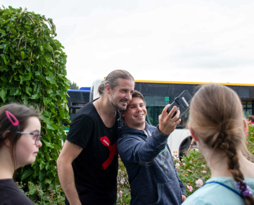 Ein Mann macht ein Selfie mit seinem Handy. Zwei Mädchen stehen im Hintergrund.