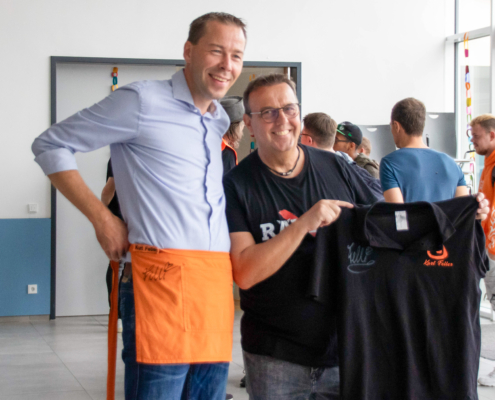 Ein Mann hält ein schwarzes T-Shirt hoch. Er trägt eine blaue Hose und eine Schürze. Im Hintergrund stehen andere Leute.