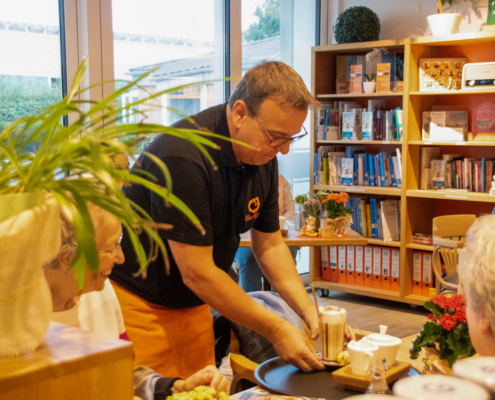 Ein Mann gibt Essen aus. Er steht in einem Raum mit Büchern. Es sitzen Leute am Tisch.