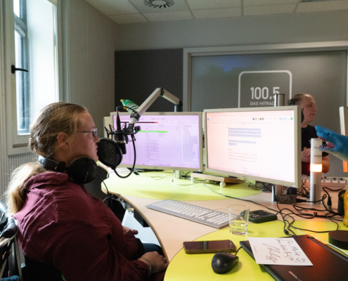 Ein Mann sitzt vor einem Mikrofon. Er spricht wahrscheinlich in ein Radio. Es gibt viele Geräte und Bildschirme auf dem Tisch.