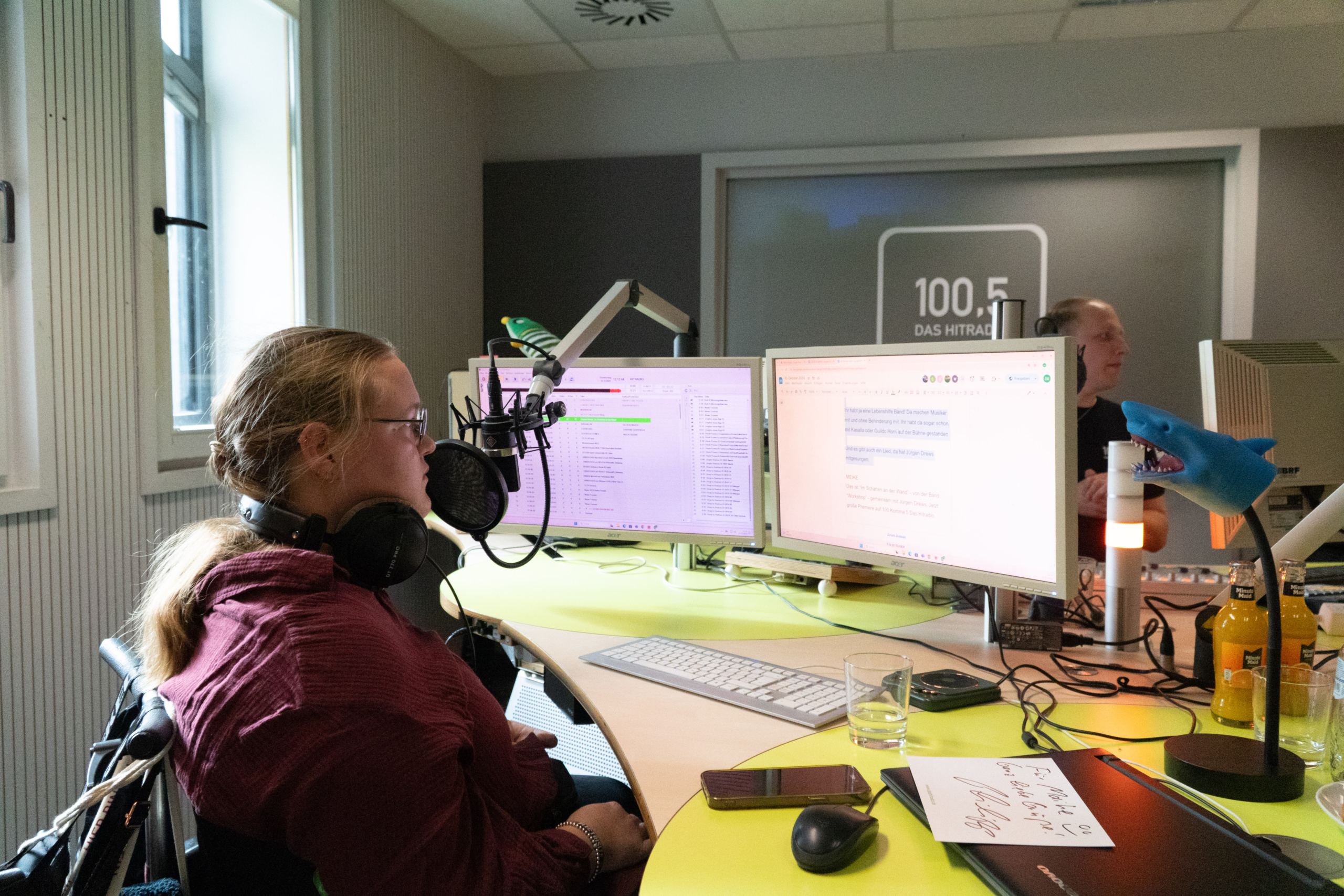 Ein Mann sitzt vor einem Mikrofon. Er spricht wahrscheinlich in ein Radio. Es gibt viele Geräte und Bildschirme auf dem Tisch.