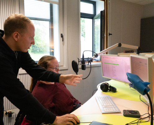 Zwei Personen sitzen vor einem Computer. Eine Person zeigt auf den Bildschirm. Es sind Mikrofone und Kopfhörer zu sehen.