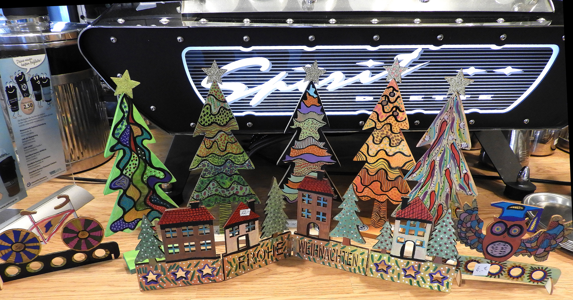 Bunte Pappfiguren von Weihnachtsbäumen und Häusern stehen auf einem Tisch. Es sieht nach einer lustigen Weihnachtsdeko aus.
