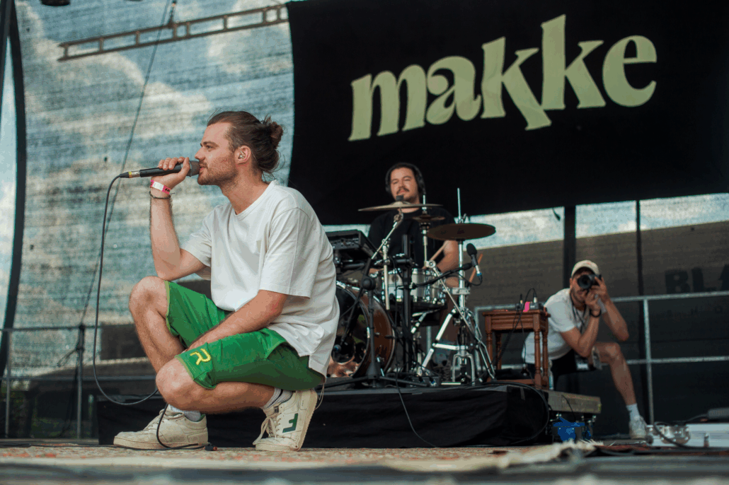 Ein Mann singt auf der Bühne. Er kniet und hält ein Mikrofon. Hinter ihm sind noch zwei Männer mit Musikinstrumenten.